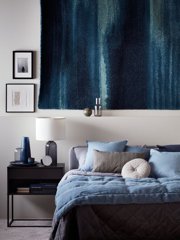 Une lampe de table TONVIS est posée sur une table de chevet VIKHAMMER noire à côté d'un lit SLATTUM avec un linge de lit KOPPARBLAD et un tapis bleu au mur.