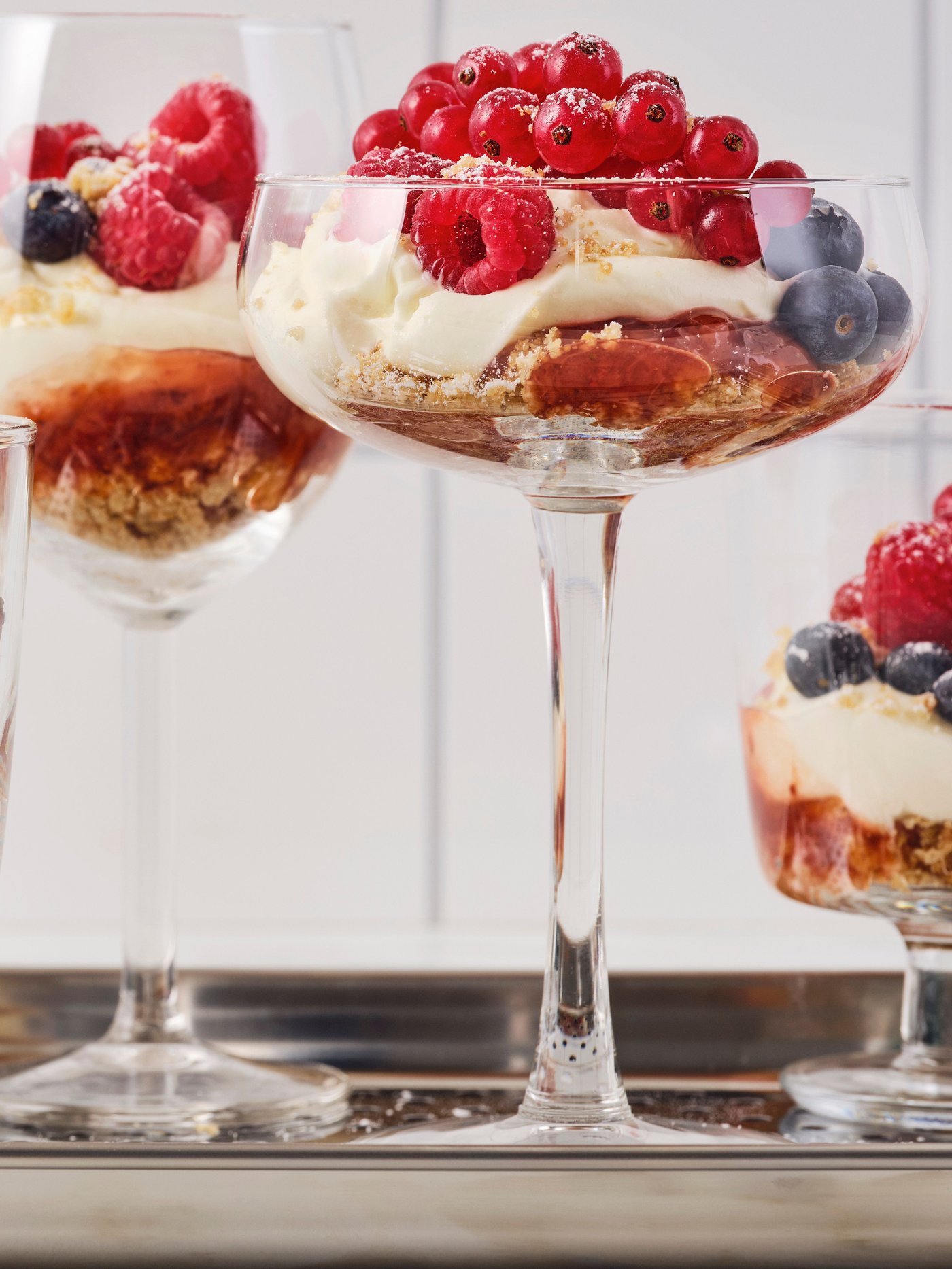 Glasses in various shapes filled with oatmeal cookies and cheesecake with raspberry jam are placed on a metal tray.