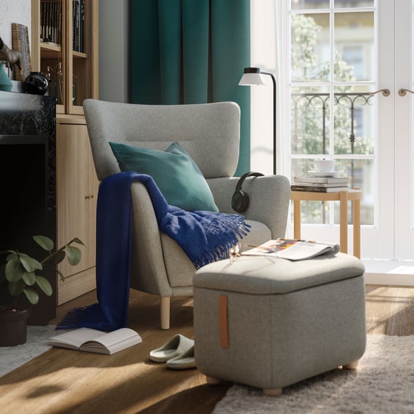 Gemütliche Leseecke mit Sessel, Fußhocker, Büchern, Decke und Kopfhörern – stilvoll eingerichtet mit Beistelltisch, Lampe und Pflanzen bei natürlichem Licht.
