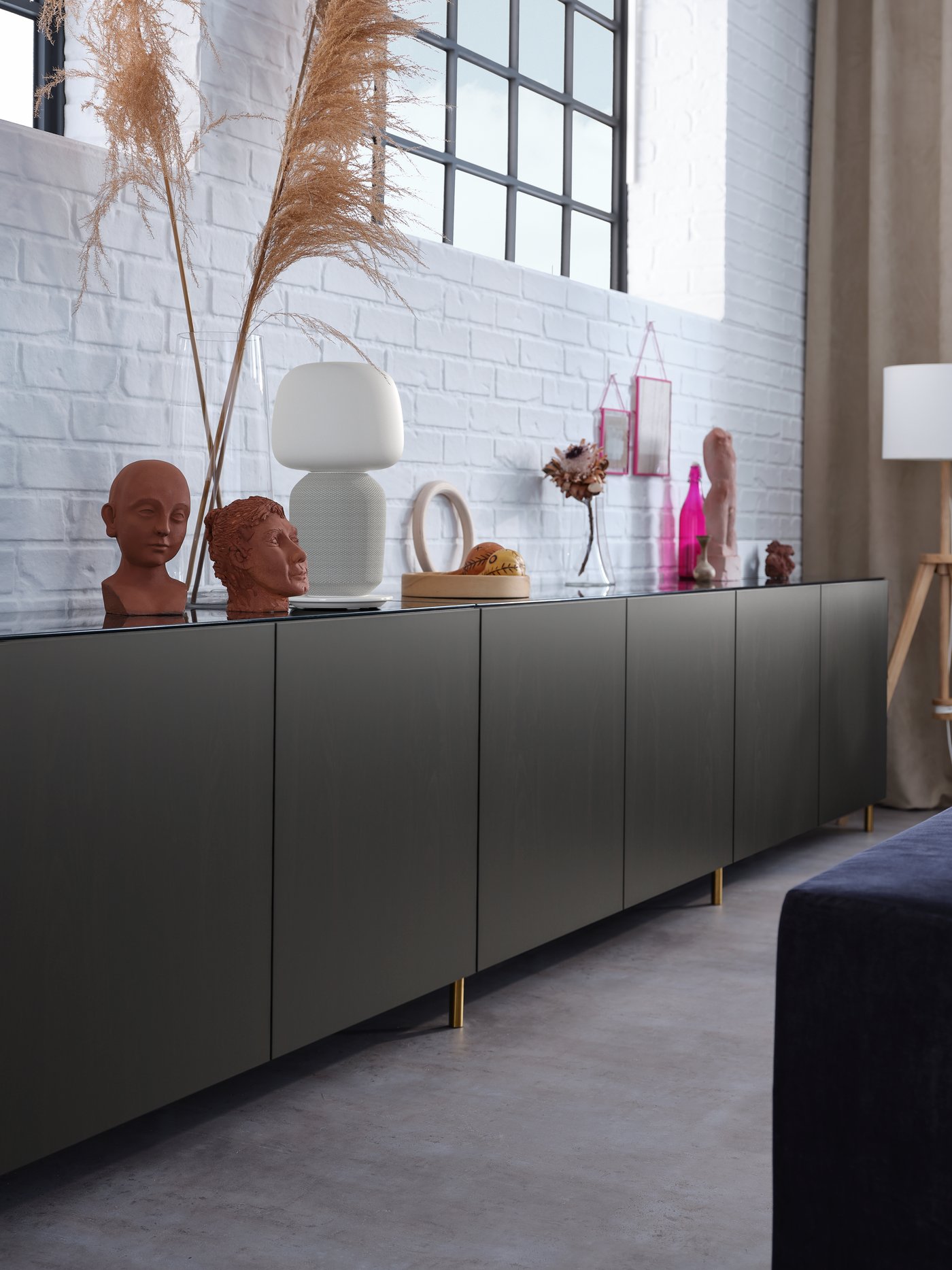 UnpopulatedEen modern interieur met een lang donker dressoir tegen een witte bakstenen muur, versierd met bustes, een witte lamp, pampasgras in een vaas en andere decoratie, naast een groot raam met zwarte omlijsting en gordijnen.