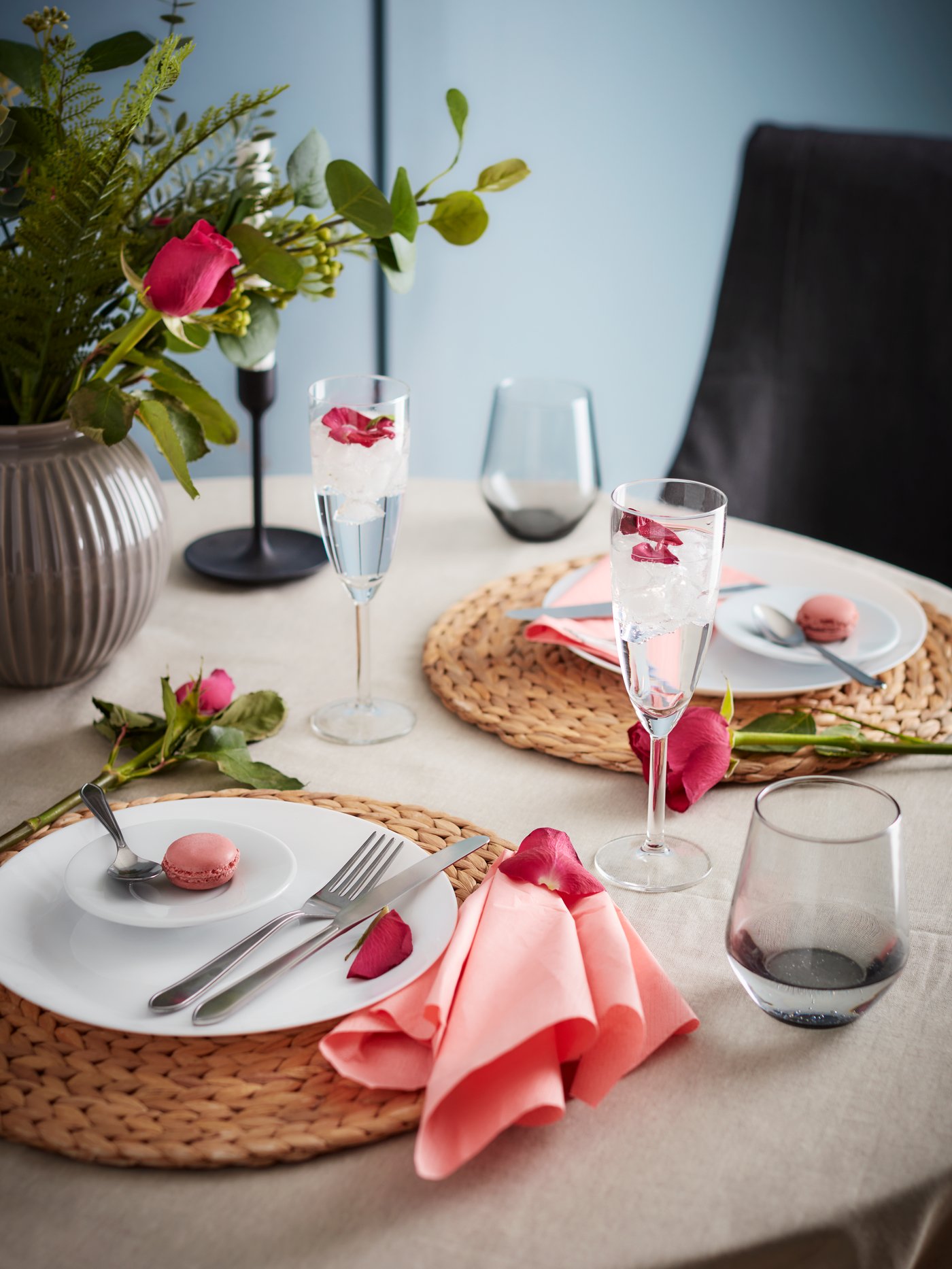 Una mesa redonda puesta con individuales redondos SOARE sobre un mantel beige, platos blancos, copas de champaña y vasos cortos. En el centro de la mesa hay un florero con rosas rojas. 