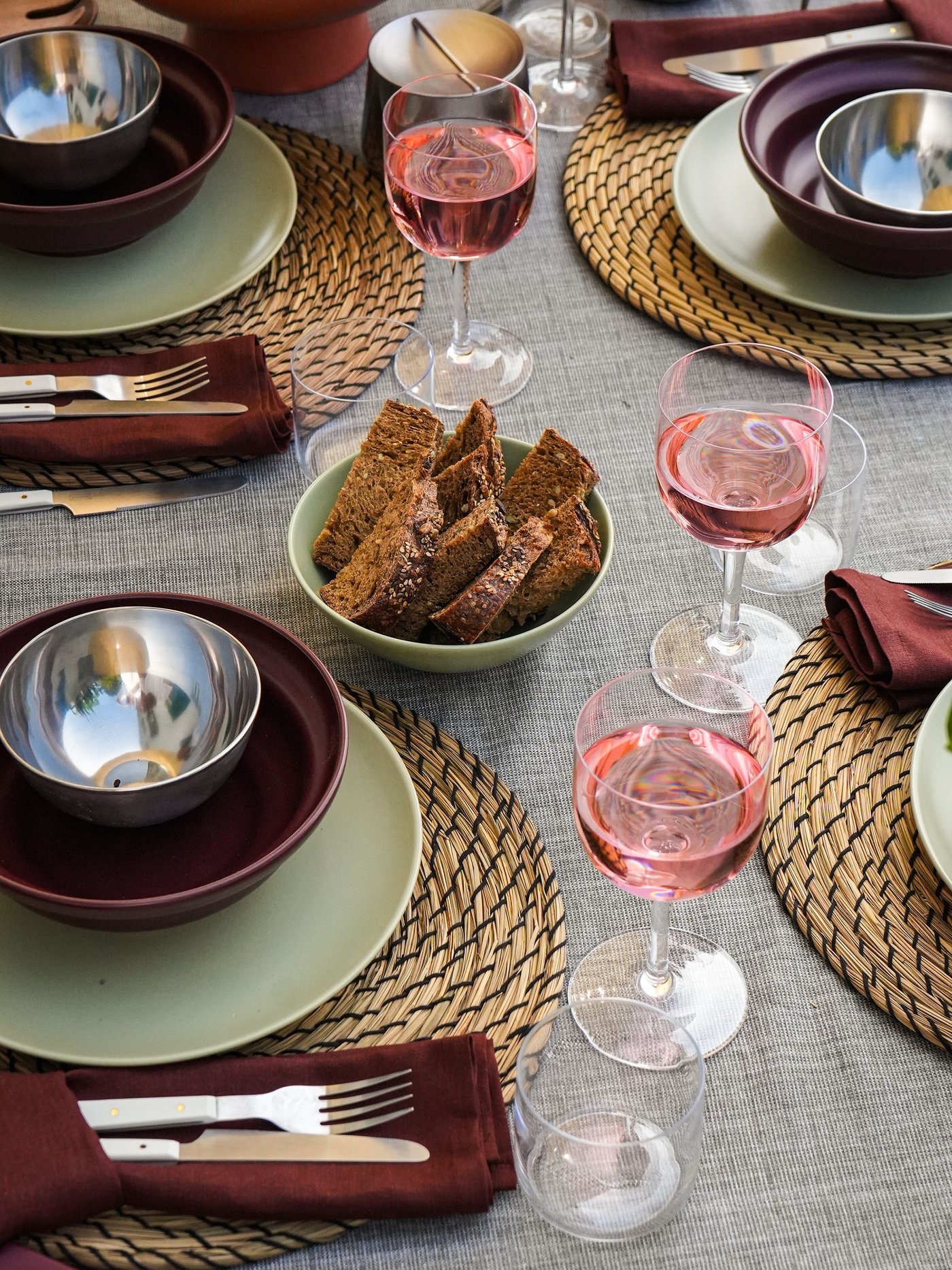 Gedeckter Tisch mit geflochtenen LÄTTAD Platzsets, hellgrünen FÄRGKLAR Tellern, rotbraunen SPJUTLILJA Schalen, Besteck und Roséwein. Brot, Käse, Oliven.