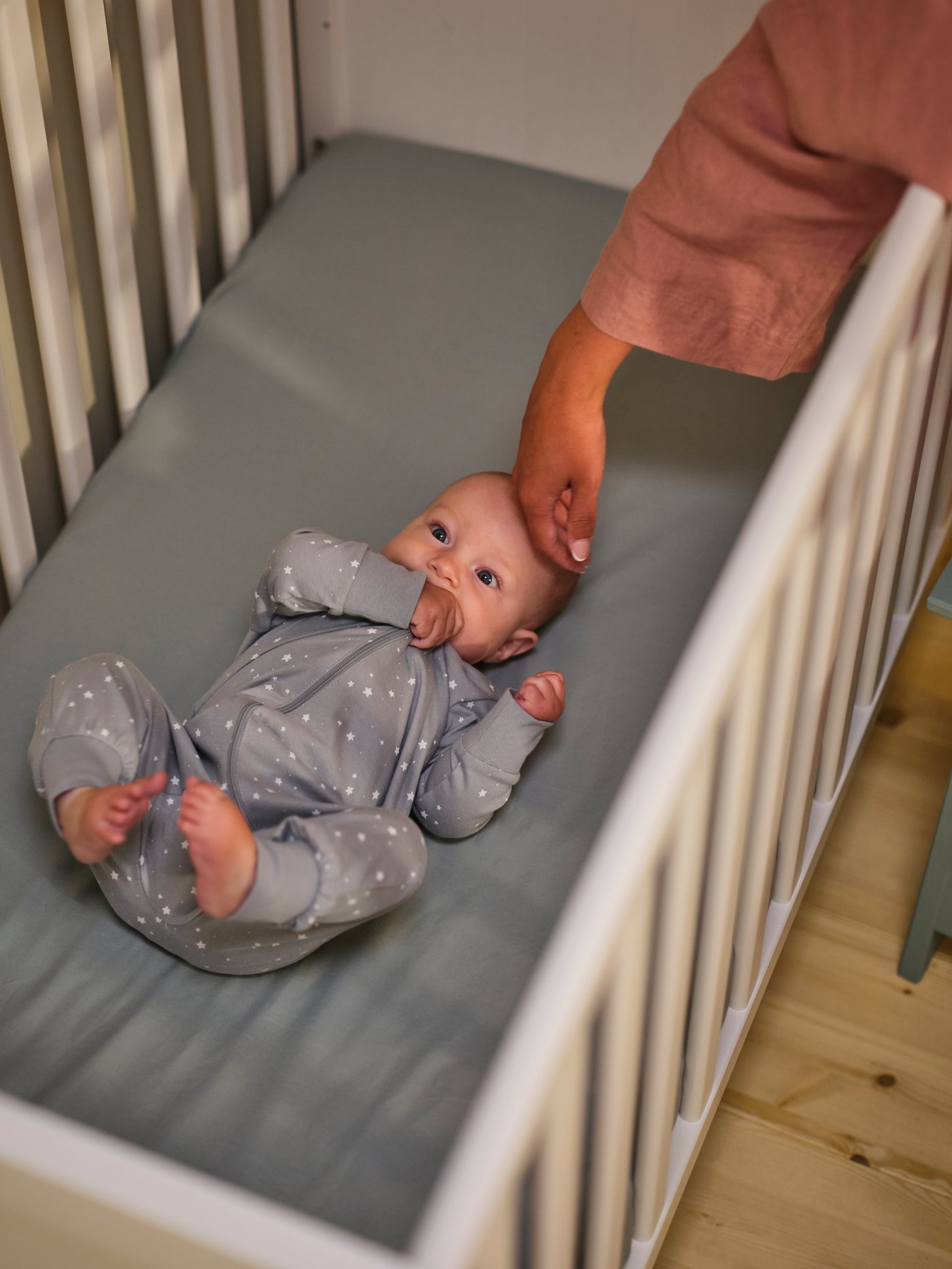 Ein Baby liegt in einem weißen SUNDVIK Gitterbett mit blauem GULDVÄVARE Leintusch auf der Matratze. Eine Hand streichelt das Baby,
