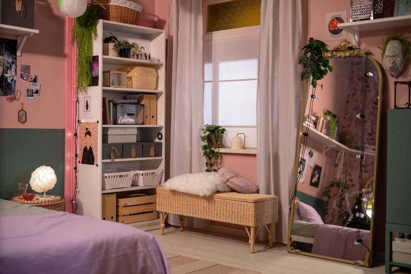 Een gezellige IKEA slaapkamerhoek met een bed, een kast en een bankje, aangevuld met een staande spiegel en planten tegen roze en groene muren.