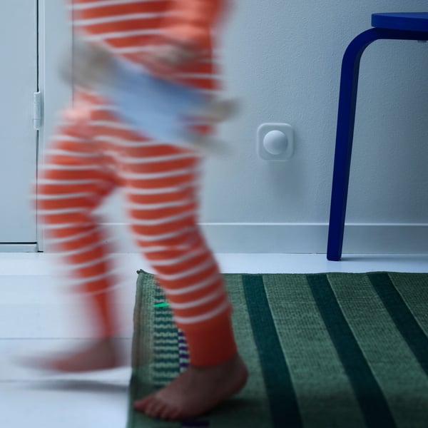 Dete u crvenoj pidžami s belim prugama ide na jednu stranu držeći igračku, u pozadini se vide plava stolica i tepih.