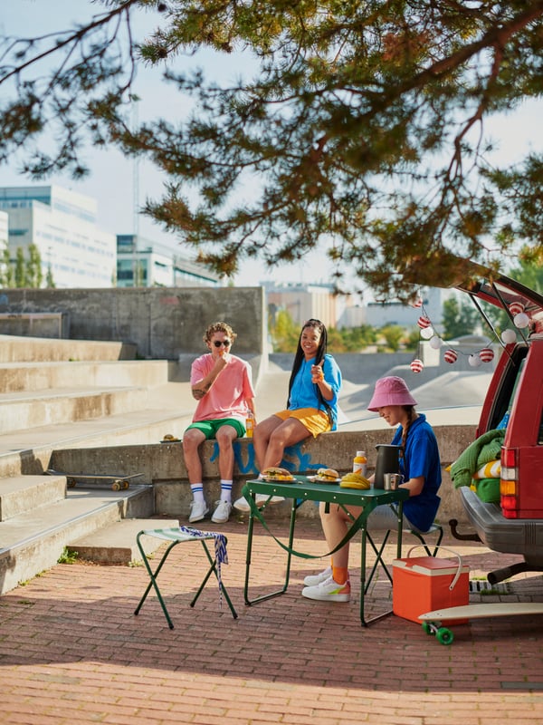 Three young people in a sunny city landscape sitting on a stone wall and at a green STRANDÖN folding table set next to a car.