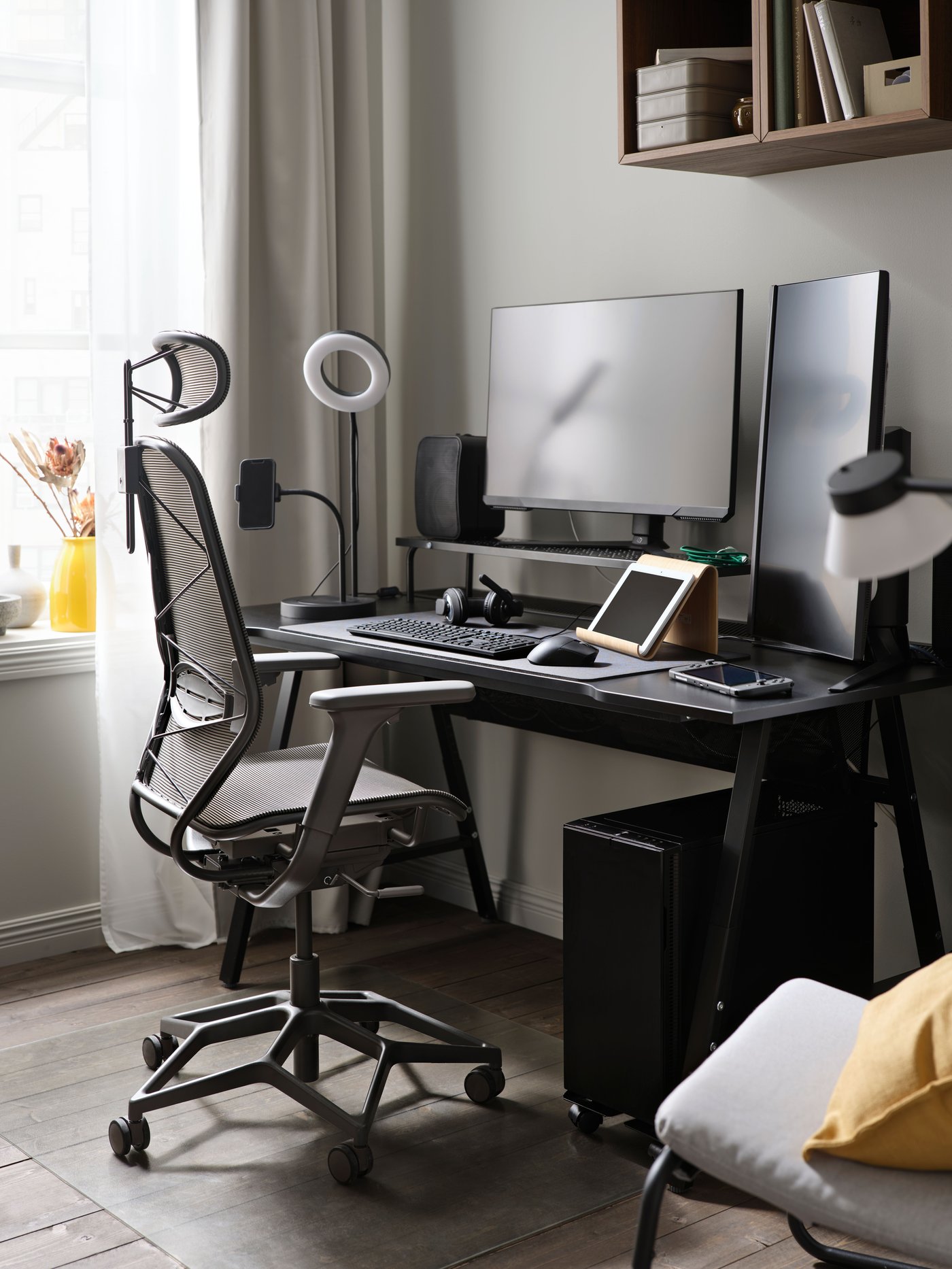 A black UTESPELARE gaming desk on a grey wall holds two monitors and accessories. A grey mesh gaming chair faces the desk.