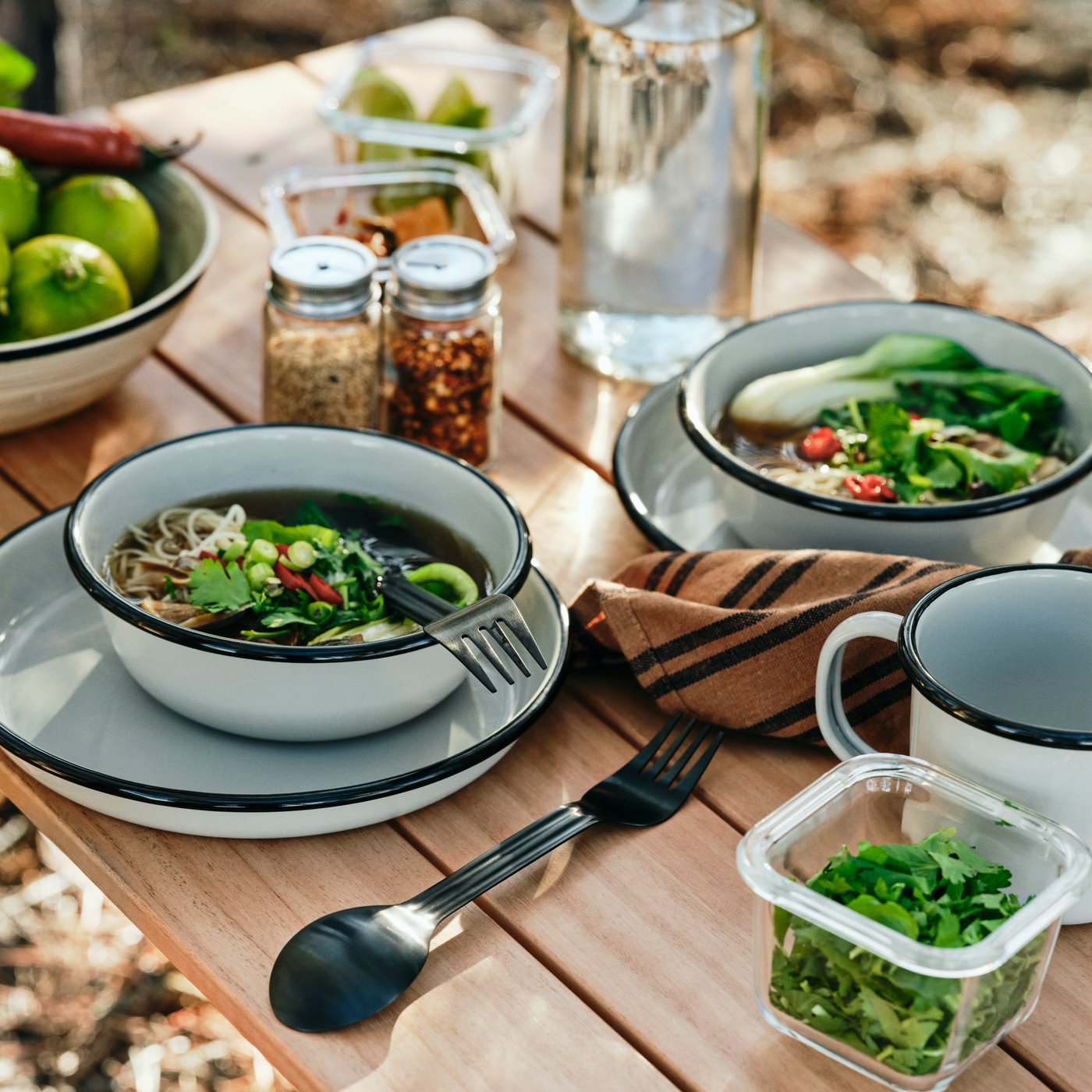 Two SOLUPPGÅNG bowls with noodle soup in them and two SOLUPPGÅNG sporks are on a folding table, along with other dining items.