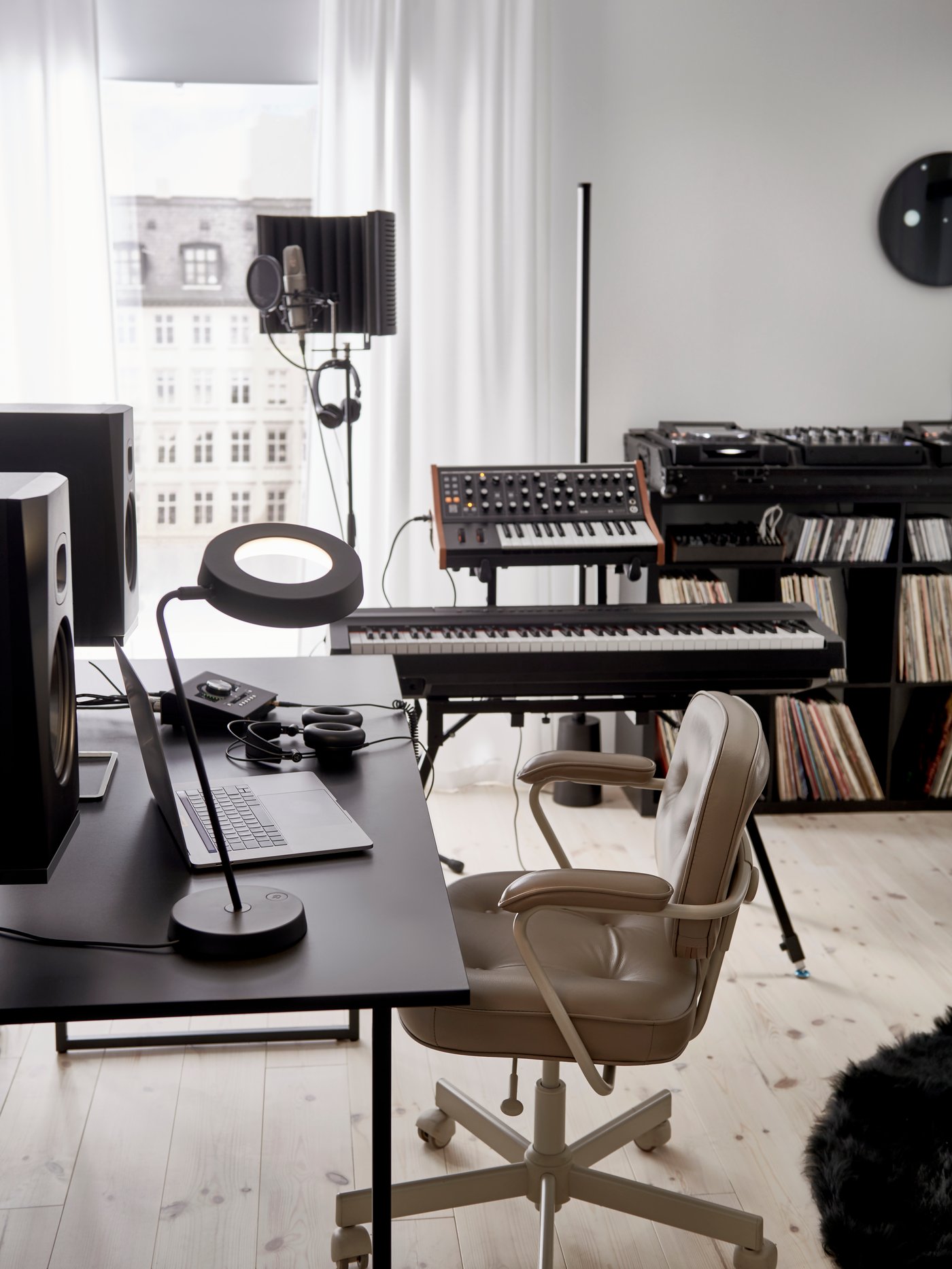 A black OBEGRÄNSAD desk, shelving unit and work lamp in a white room with speakers, keyboards, mic and decks by a window.