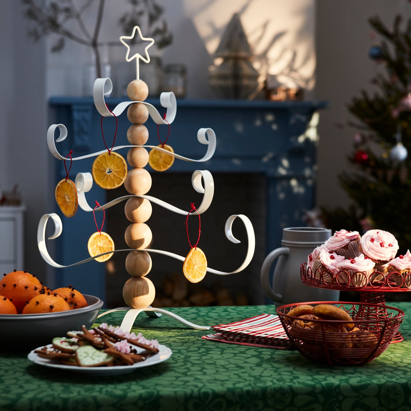 Orange peels are hung as tree decorations on an artificial Christmas tree. The artificial tree made of metal and wood stands on a table with green tablecloth.