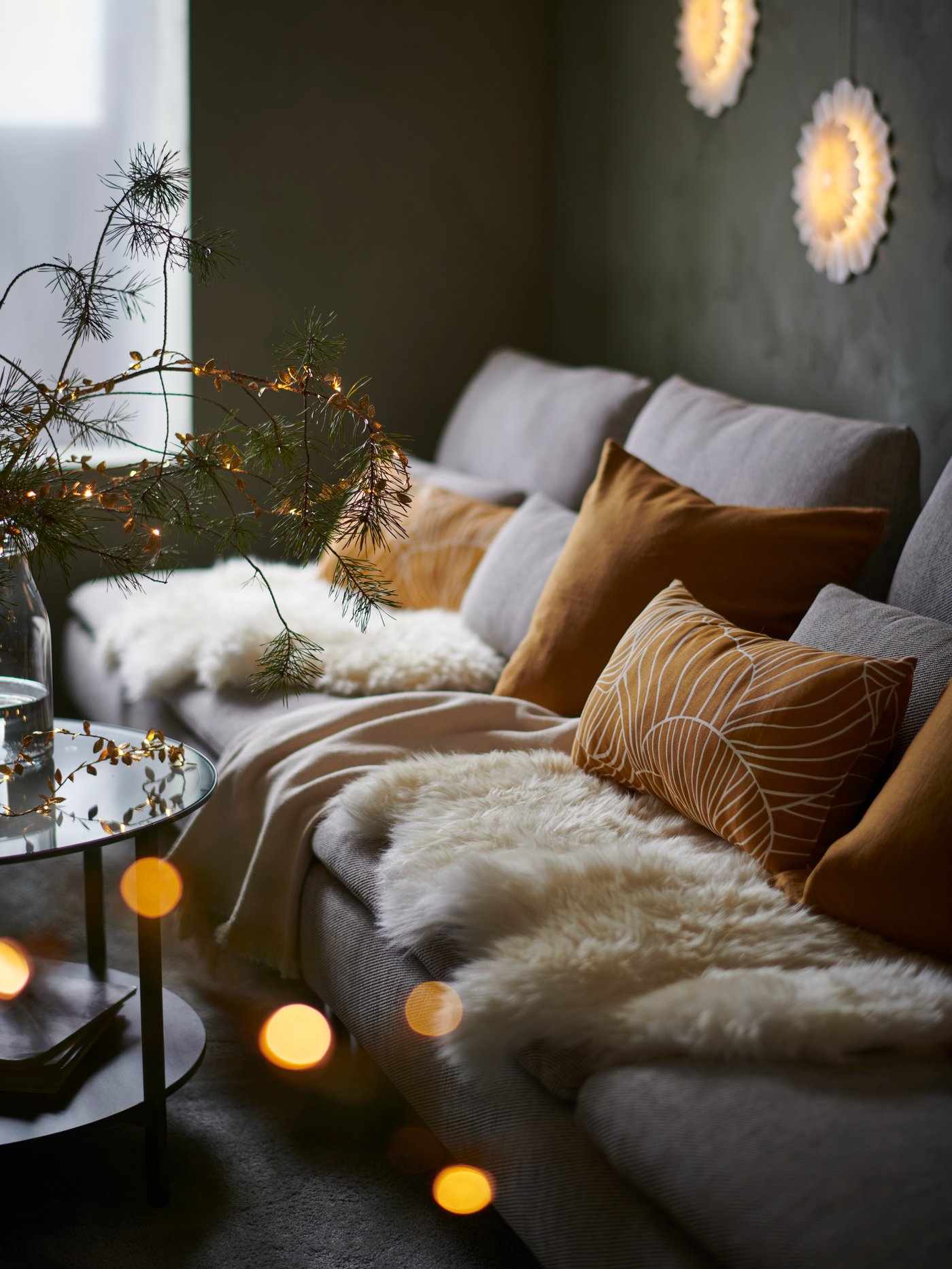 A long sofa with golden and white decorations in and around it, including SVAMPMAL cushions and STRÅLA LED lighting chains.
