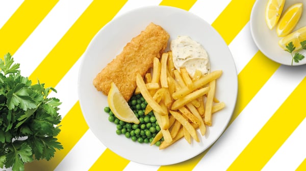 Knuspriges Fish-&-Chips-Menü mit Pommes frites, Erbsen, Zitronenspalte und Remouladensauce auf einem weißen Teller vor gelb-weiß gestreiftem Hintergrund.