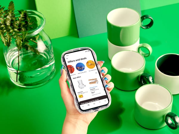A hand with green-painted fingernails is holding a smartphone displaying the IKEA app over a green table with mugs.