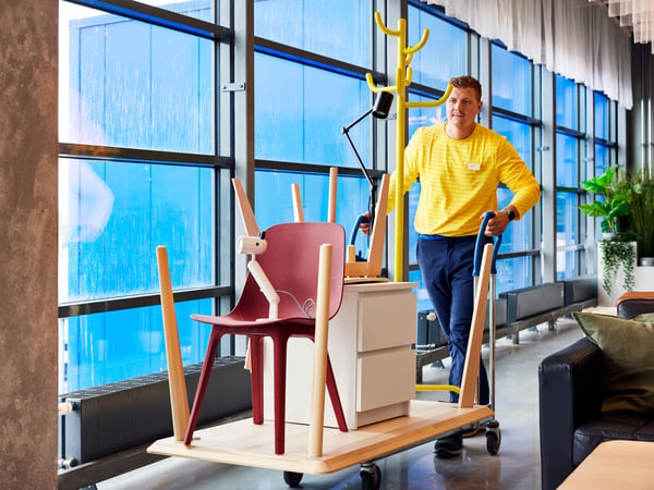 Zaposleni u kompaniji IKEA u žutoj košulji gura kolica puna kancelarijskog nameštaja, uključujući crvenu stolicu i bele fioke, kroz moderni hodnik sa staklenim panelima.