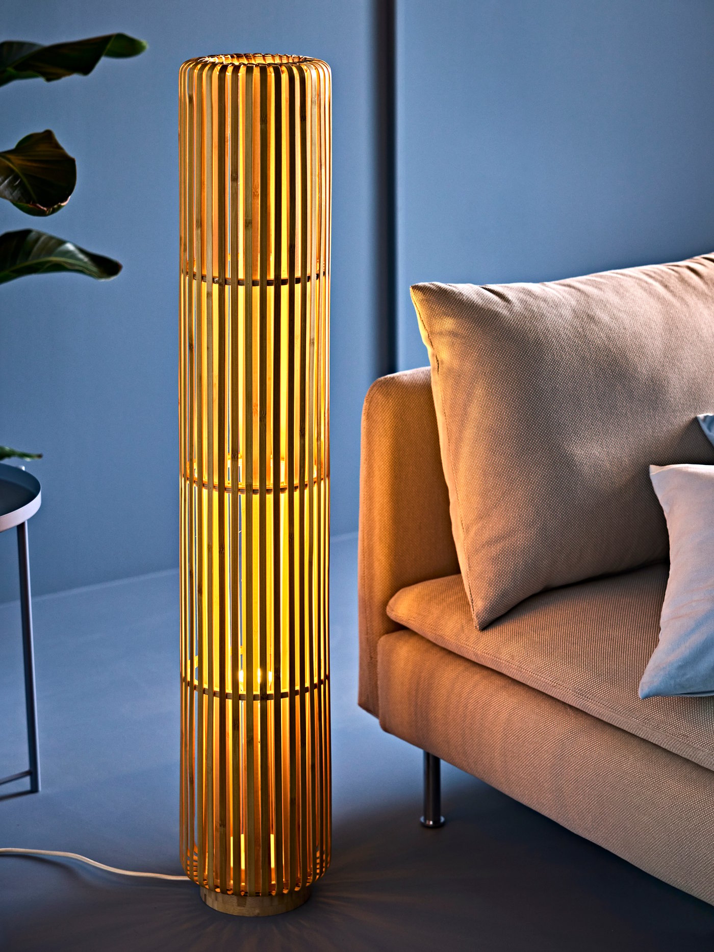 A living room with a VARPTROSS bamboo floor lamp casting a warm glow, beside a beige sofa with cushion and pillows.