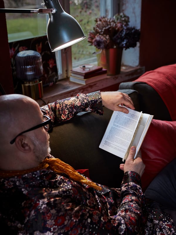 In a cozy living room, a man lounges on a sofa while reading a book. A floor lamp provides lighting as he reads.