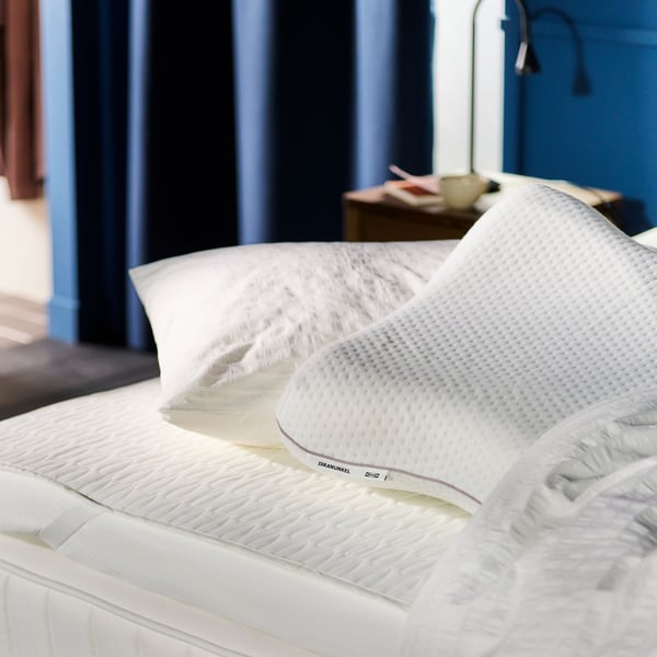 An unmade bed with white bedding and ergonomic pillows in a pile on top of it. Blue curtains in the background.