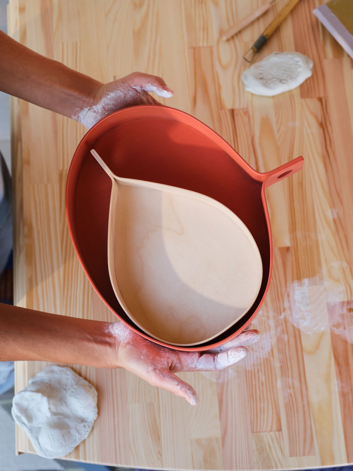Two flour-dusted hands hold an orange birch veneer OMMJÄNGE tray with a smaller birch veneer OMMJÄNGE tray inside it.