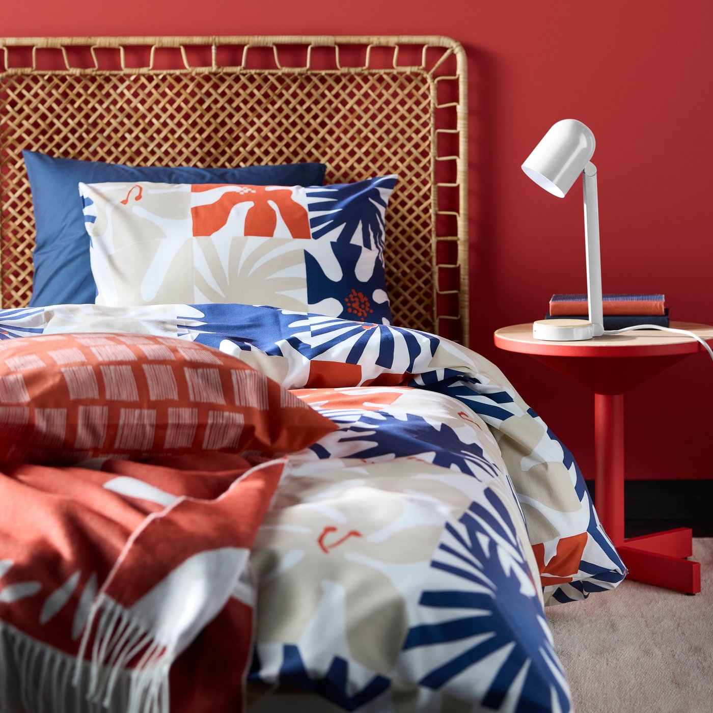 A white desk lamp sitting on a red side table placed beside a bed with a rattan headboard, made with HYLSNEJLIKA bed linen.