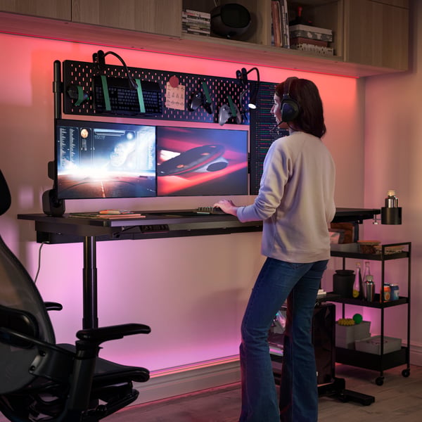 A person wearing a headset is standing and gaming at a MÅLOMRÅDE gaming desk. A NISSAFORS trolley in black is by the desk.