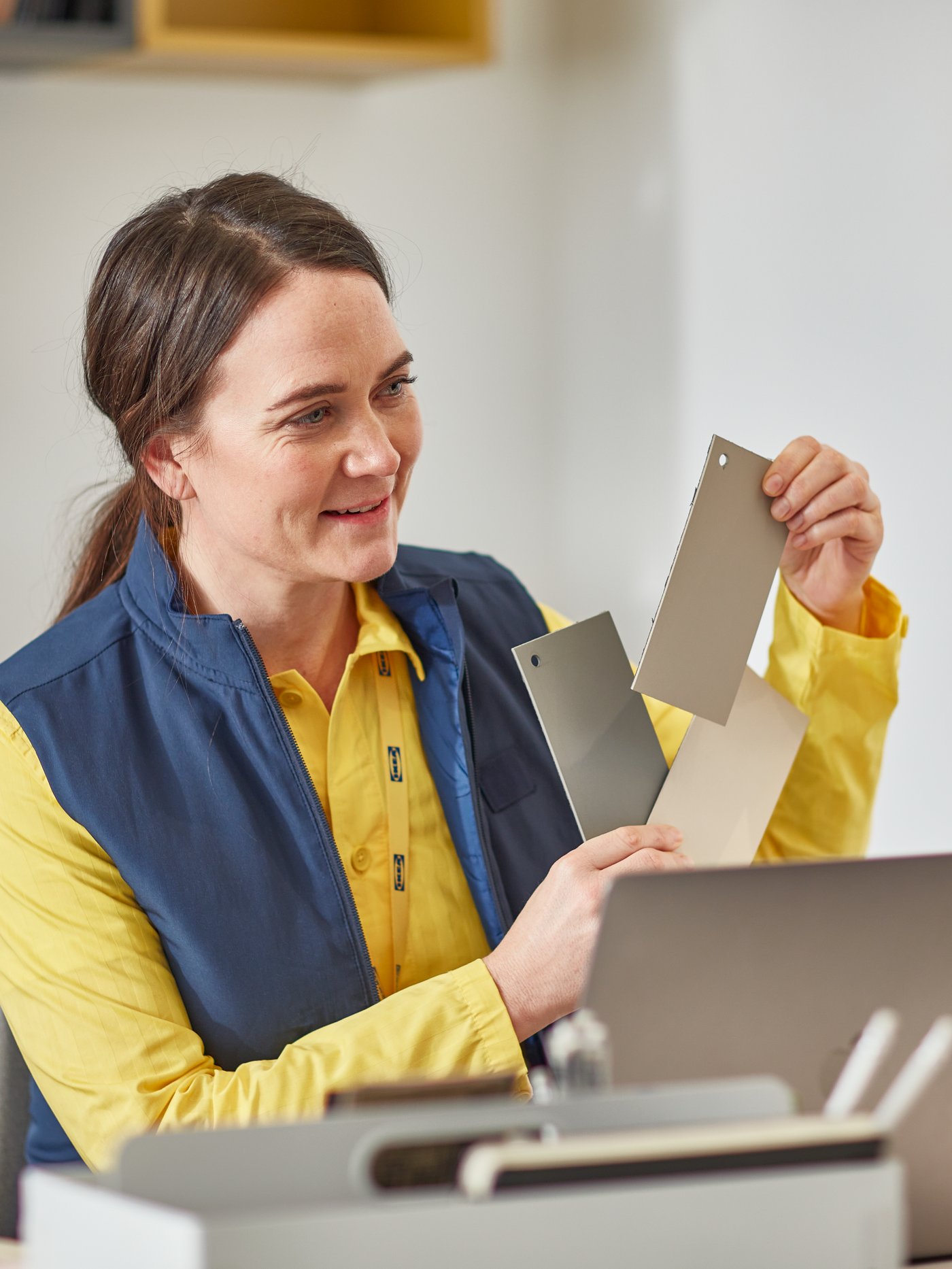 An IKEA co-worker in a yellow shirt sits behind a laptop and holds up three colour samples in beige and brown shades.