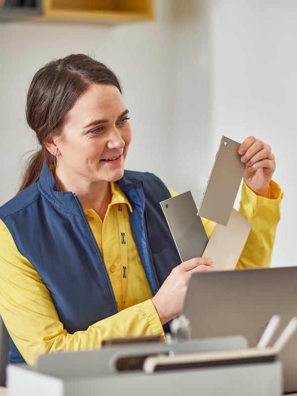 A woman in blue-and-yellow IKEA co-worker clothes holds up colour samples while looking across the desk she’s sitting at.