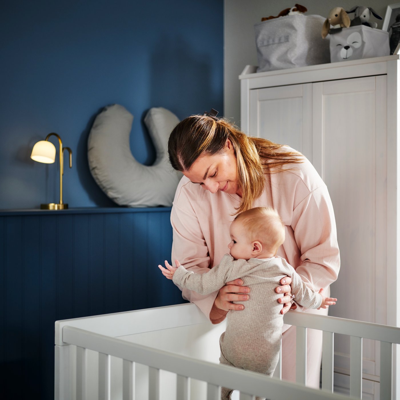 A mother picks up a baby from a white SUNDVIK cot in the corner of a dark blue bedroom with a white wardrobe behind them.