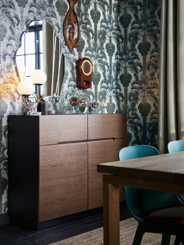 Ein dunkles Sideboard mit einem Spielgel darüber an einer Wand mit Mustertapete aus floralen Ornamenten