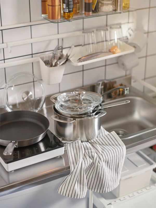 HEMKOMST cookware sits on a SUNNERSTA mini kitchen, with a tea towel. Glasses and cutlery hang behind it on a wall rack.