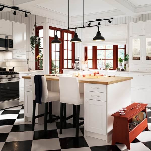 Une cuisine lumineuse avec un sol noir et blanc comporte un grand îlot de cuisine avec des façades AXSTAD blanc mat et deux chaises hautes.