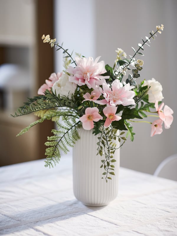 Ein SMYCKA Kunstblumenstrauss und einige SMYCKA Kunstblumen stehen in einer weissen STILREN Vase auf einer weissen Fläche.