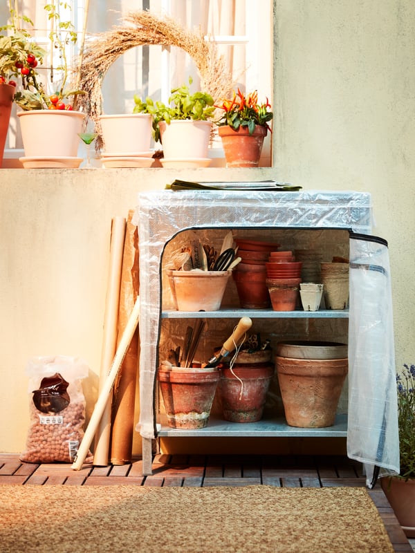 Uno scaffale in metallo con fodera ha al suo interno dei vasi in terracotta impilati e utensili da giardino. Su un davanzale sono appoggiate delle piante in vaso - IKEA