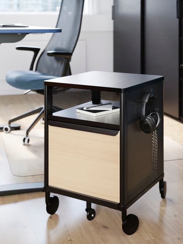 A black BEKANT storage cabinet on castors with a notebook and headphones on it, in front of a blue office chair.