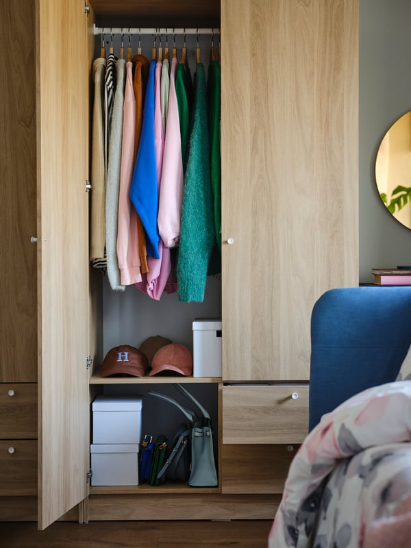 An oak-effect VILHATTEN wardrobe with an open door showing colourful clothes on hangers and two shelves with hats and boxes.