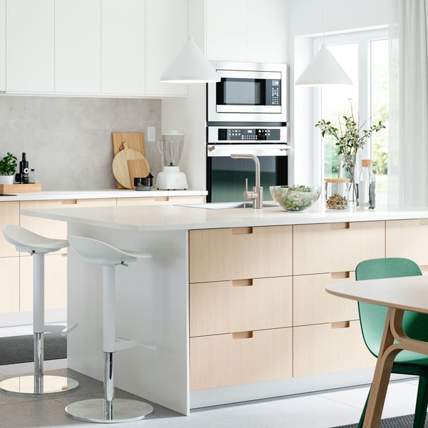 Two white JANINGE bar stools stand at one end of a kitchen island with a white top with a sink in it.