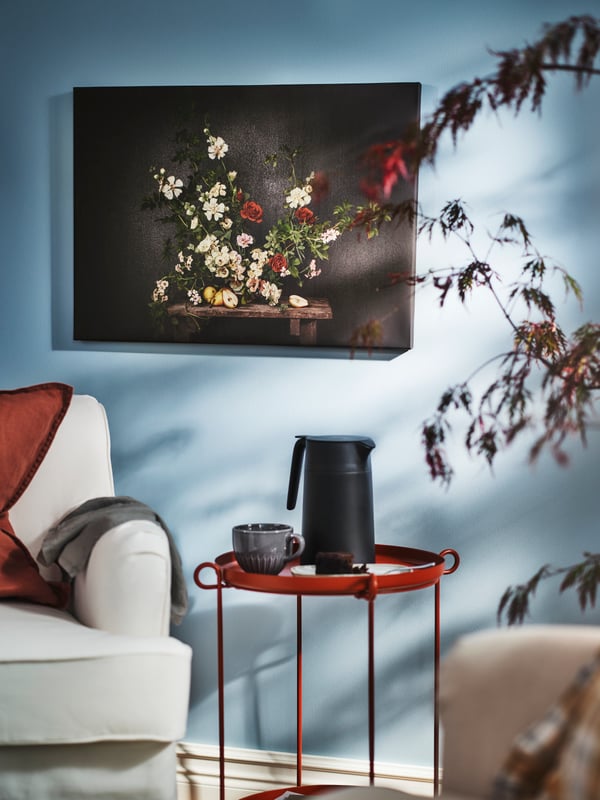 Une reproduction PJÄTTERYD représentant des fleurs sur un mur noir est accrochée au mur au-dessus d’une table-plateau LIVELYCKE sur laquelle est posée une bouteille isotherme.