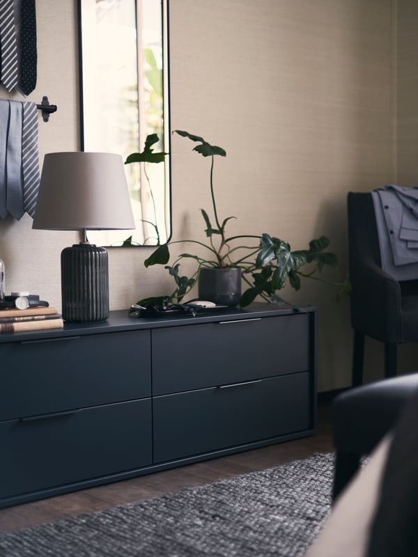 Une commode noire avec 4 tiroirs avec une plante et une lampe de table SNÖBYAR dessus. Un portant de cravates et un miroir sont fixés au mur.