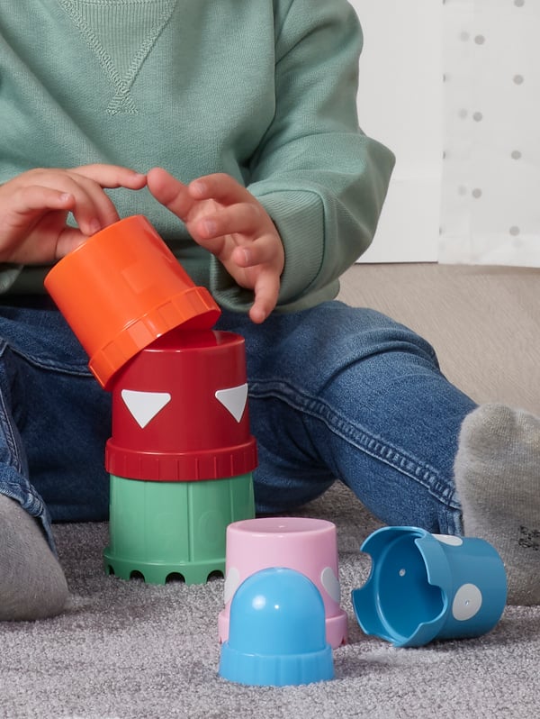 Un enfant vêtu d’un jeans bleu et d’un chandail vert est assis sur un tapis gris. Il joue avec des gobelets empilables UPPSTÅ multicolores.