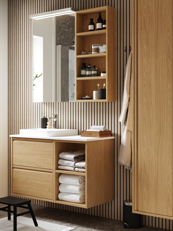 An ÄNGSJÖN washstand in oak effect with drawers and open shelves is placed on a bathroom wall with a mirror cabinet above it.