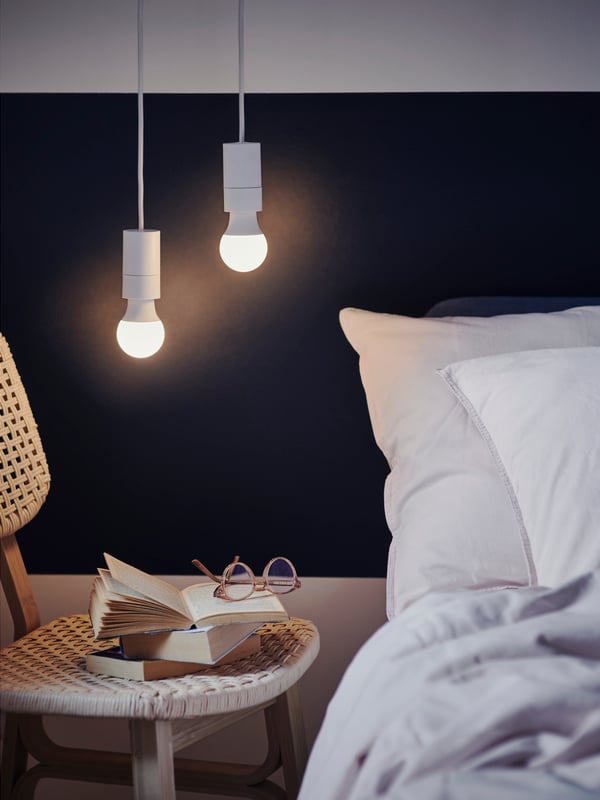 Two different SOLHETTA LED bulbs in SUNNEBY cord sets hang down over a light bamboo VOXLÖV chair next to a bed.