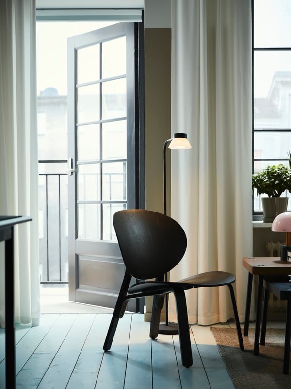 Lampadaire/liseuse SAMBORD à côté d’un fauteuil relax FRÖSET en chêne plaqué teinté en noir et d’une porte ouverte sur un balcon.