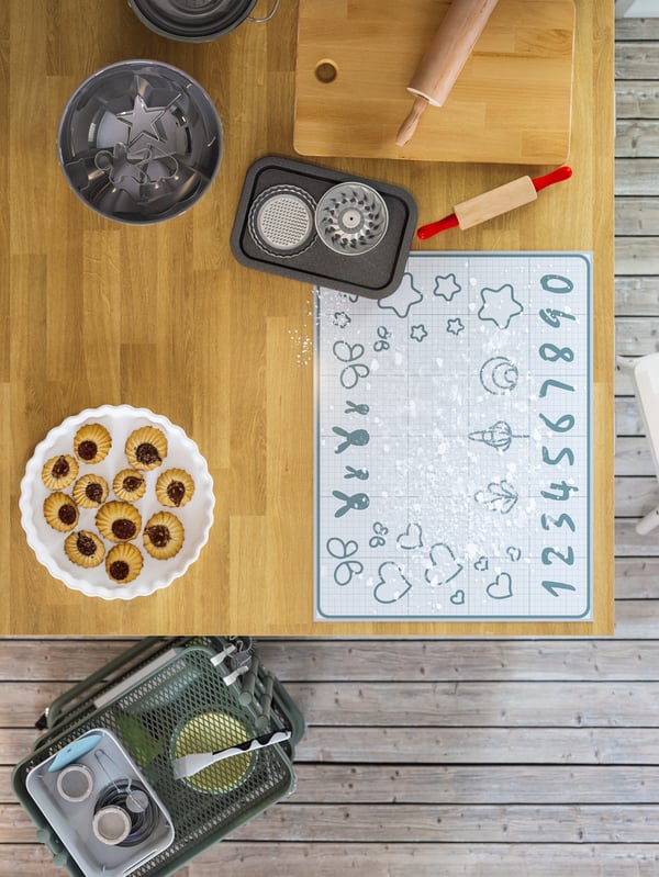 A wooden kitchen island with a baking mat to trace cookie shapes, cookies, cutting board and bakeware.