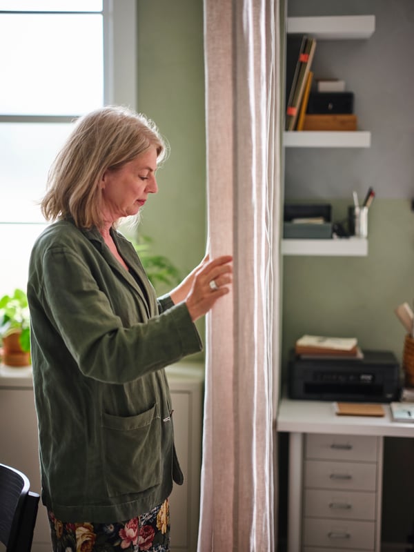 Vrouw trekt een stoffen gordijn toe dat een thuiskantoor met LACK open planken scheidt van een ander gedeelte.
