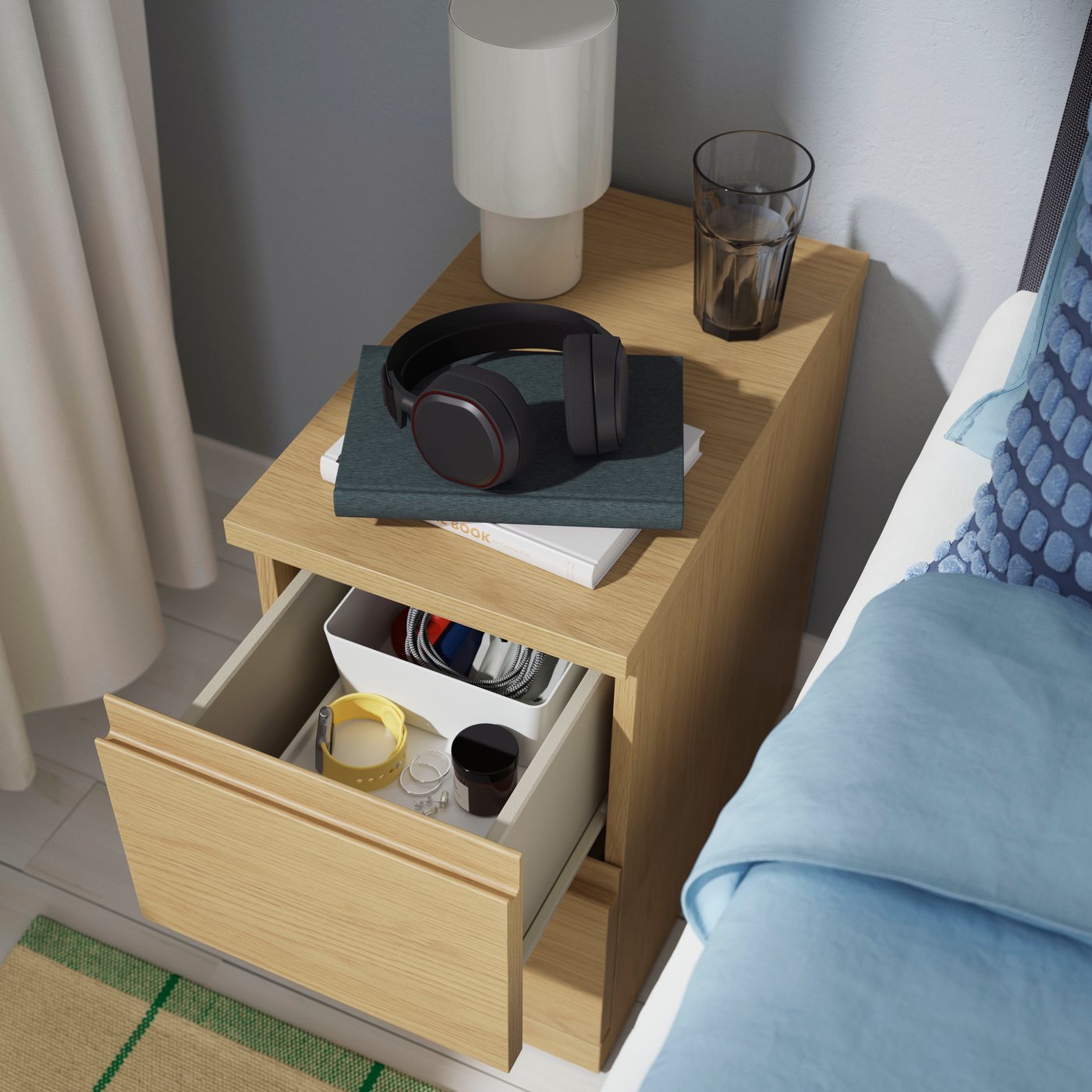 A light oak STORKLINTA bedside table with an open drawer stands next to a bed in a bedroom.