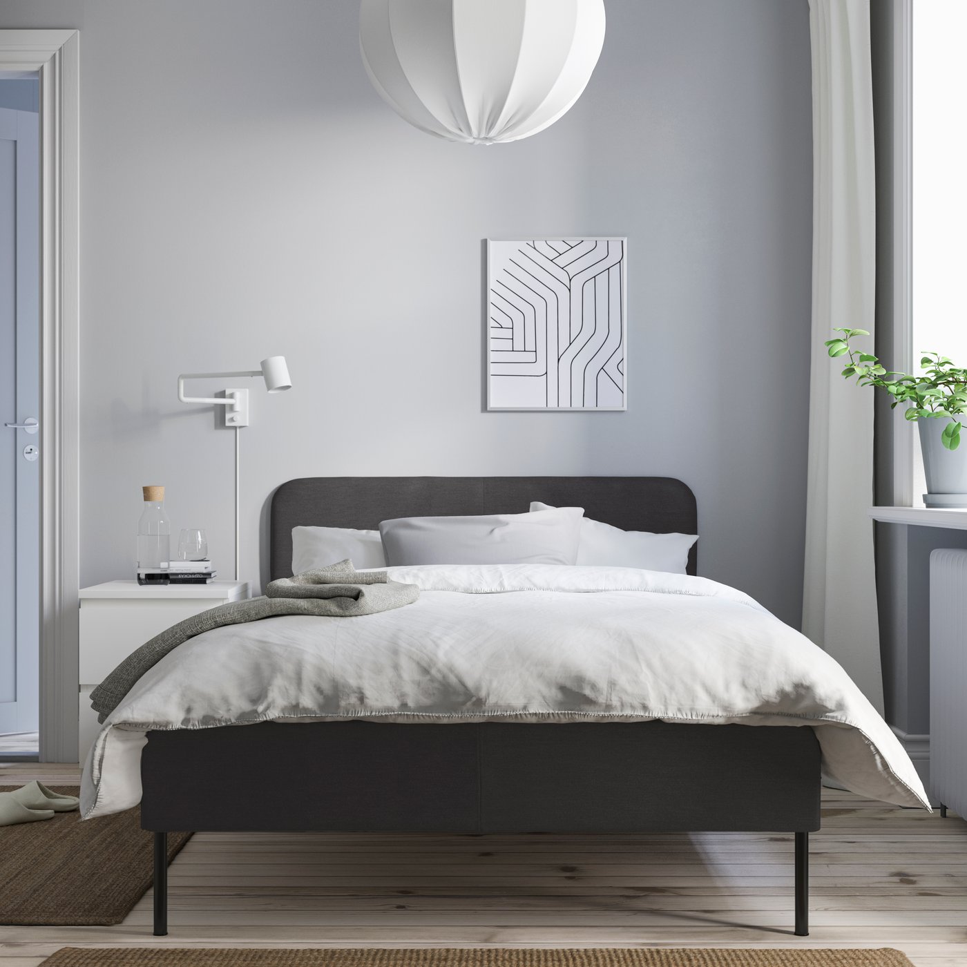 Minimalist bedroom with a dark bed frame, white bedding and soft light from a window plant.