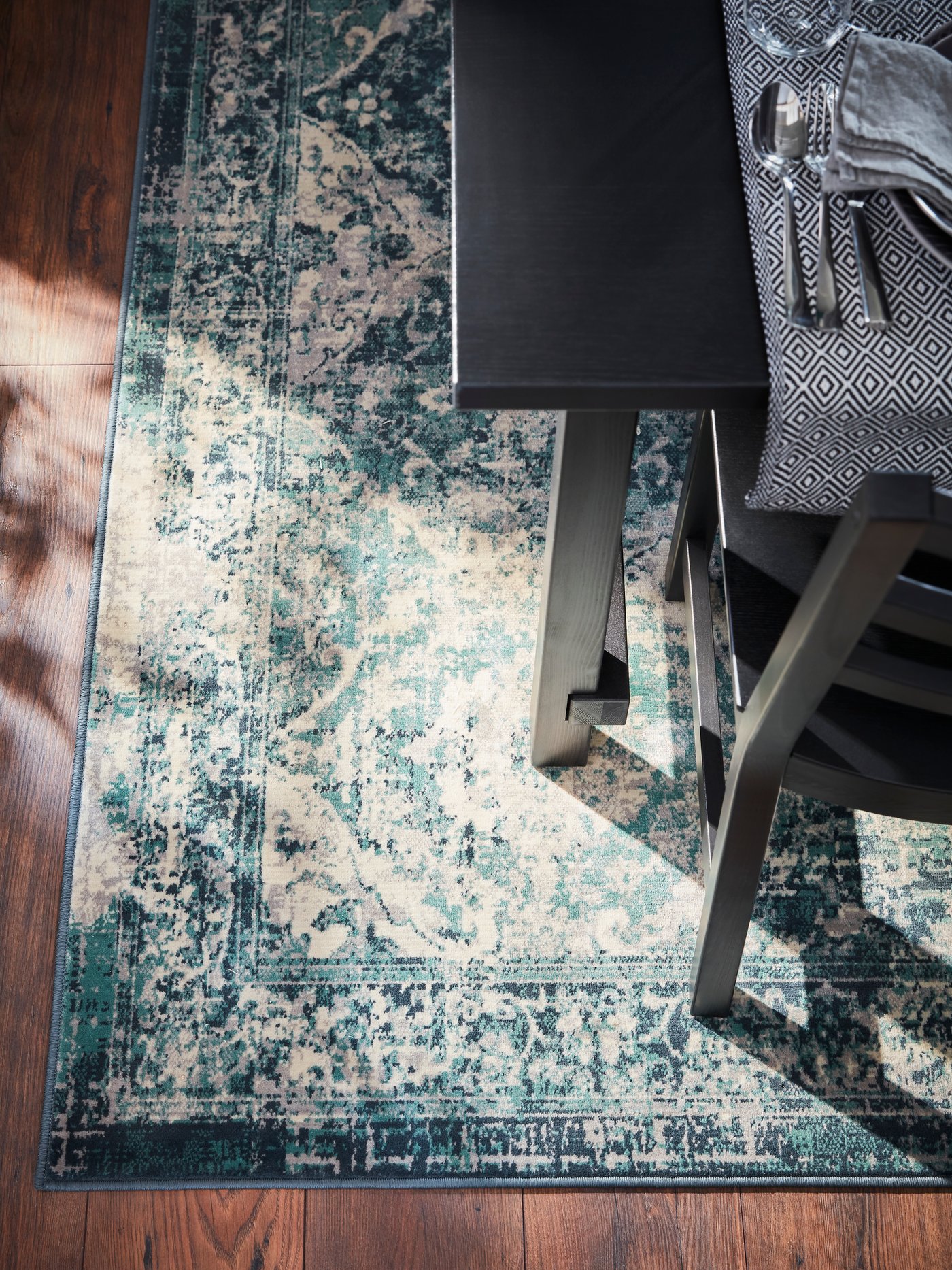 A table and a chair stand on a green/oriental-patterned rug. Cutlery and a black/white table-runner are on the table.