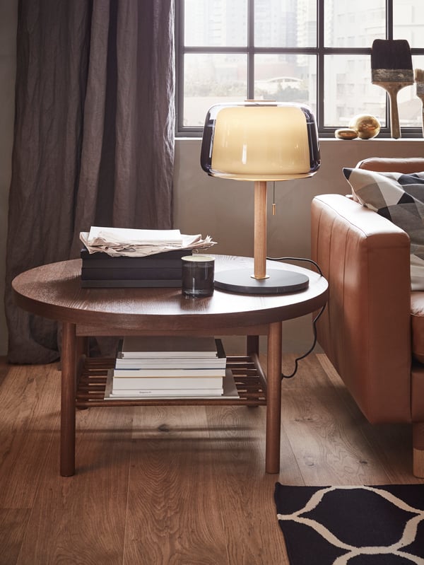 LISTERBY round dark wooden coffee table with a lamp and papers on top by the sofa.