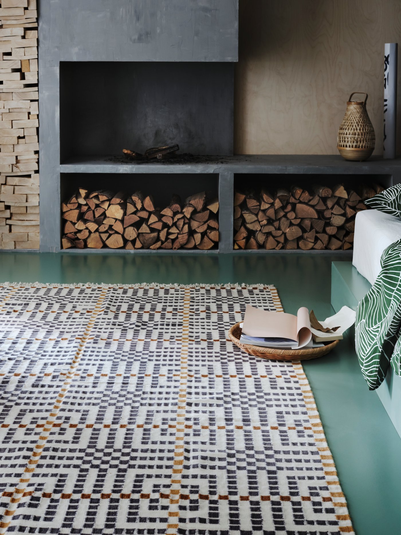 A flatwoven VAMDRUP rug with a multicoloured pattern is on a green floor in front of a rustic fireplace with logs.