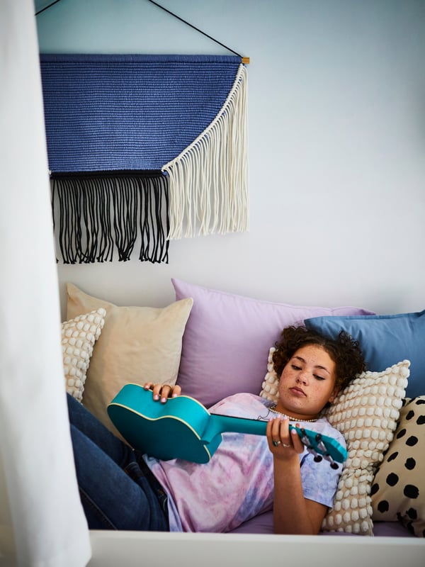 A teen lies on a bed holding a ukulele, leaning against cushions in SVARTPOPPEL and SANELA covers and NATTSVÄRMARE bed linen.