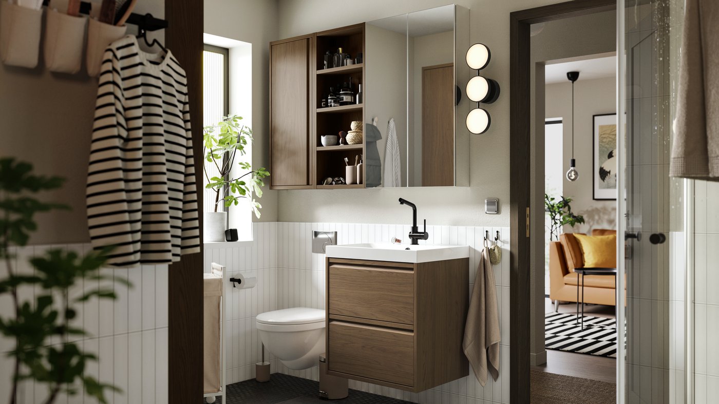 A bathroom with an ÄNGSJÖN wash-stand with drawers and wall cabinet with door in a brown oak effect seen from another room.